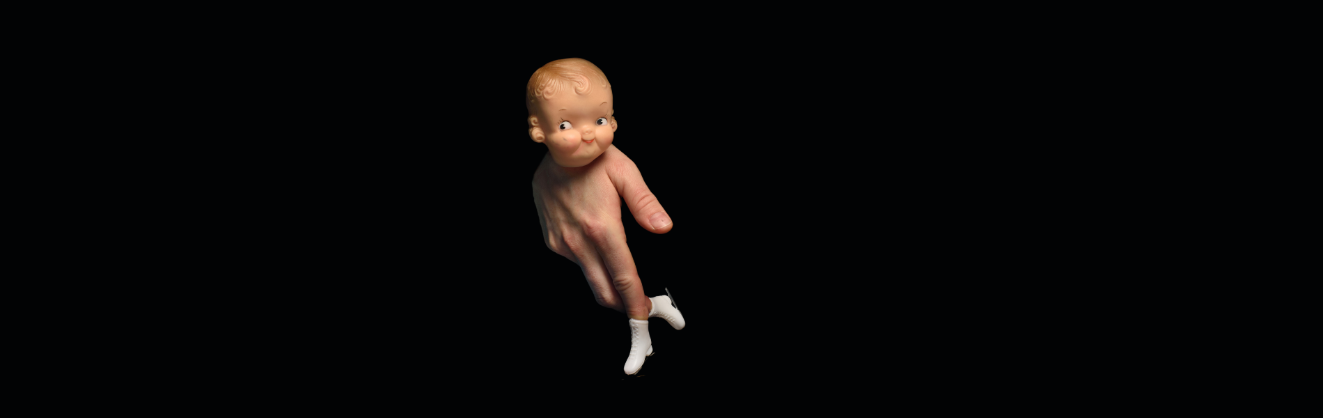 A doll head at the top of a person's hand, shaped to look like the body and legs of the doll with tiny skates on the end of the fingers, against a black backdrop