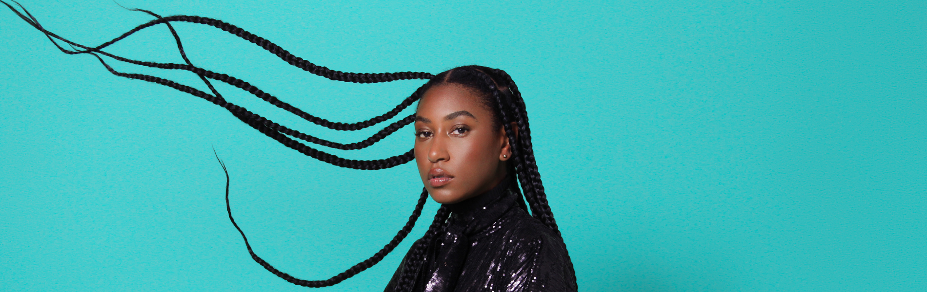 Dominique Fils-Aimé in front of a teal blue background with her hair in braids thrown out to the side of the photo