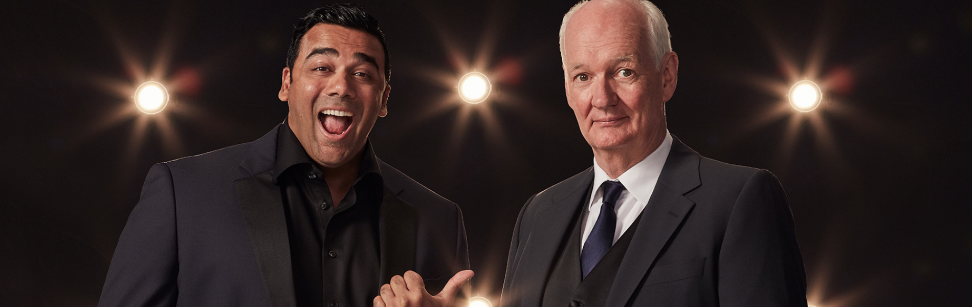 Asad Mecci and Colin Mochrie in black suit jackets standing against a black background with stage spotlights