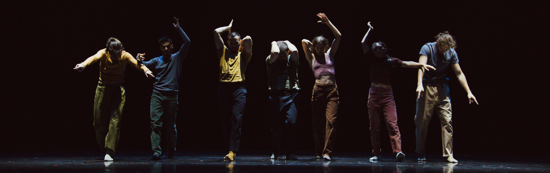 7 contemporary dancers in a line, darkly lit against a black background