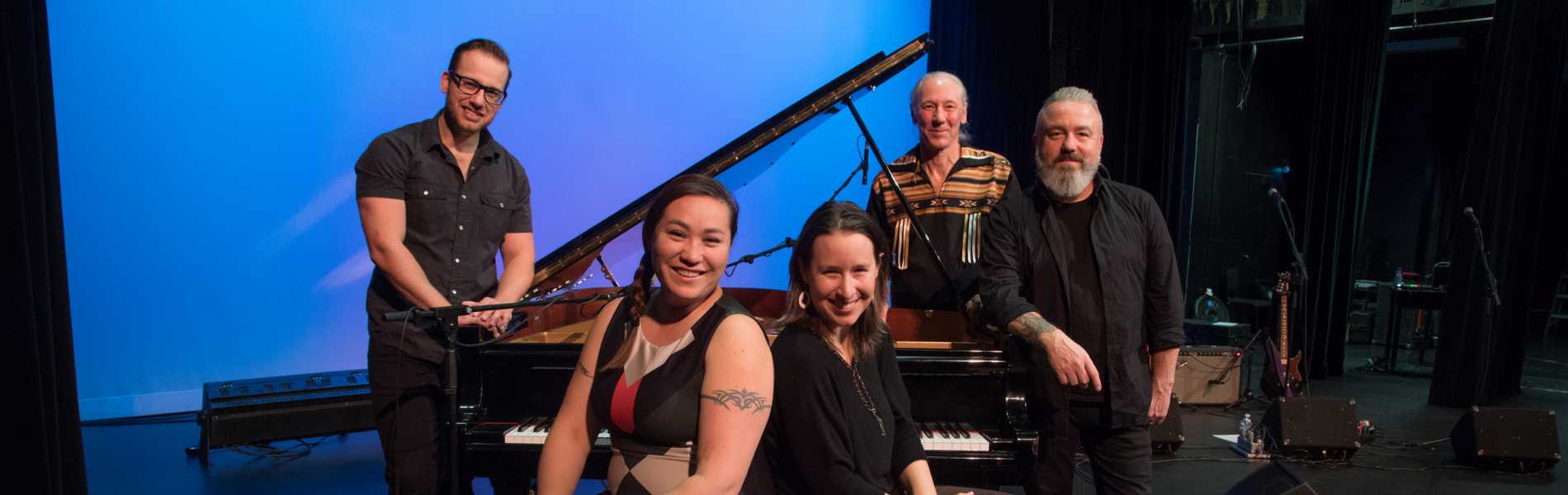 5 members of New North Collective sitting in front of a piano on stage against a blue background