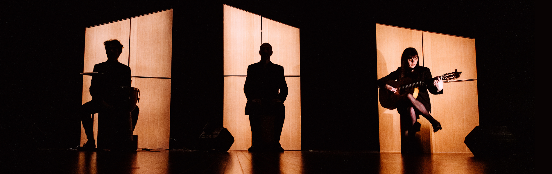 Three artists silhouetted on stage against orange window lights