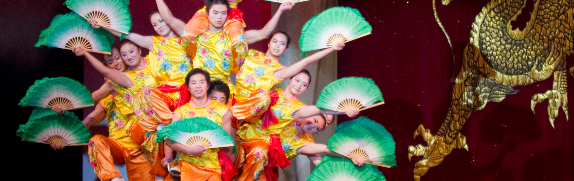 The Peking Acrobats holding large green fans out to the side as all 10 balance on a single bike