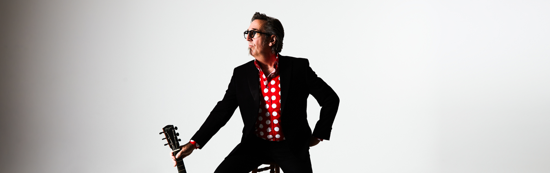 Stephen Fearing in a black jacket and red checked shirt sitting on a stool, holding his guitar out to the side