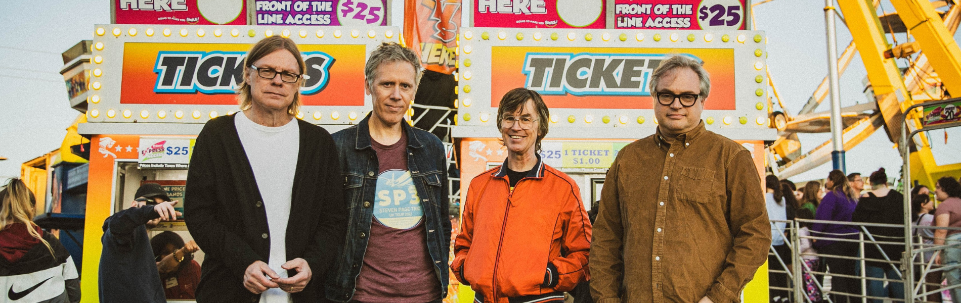 The 4 members of Trans-Canada Highwaymen looking at the camera, standing in front of a colourful carnival ticket booth