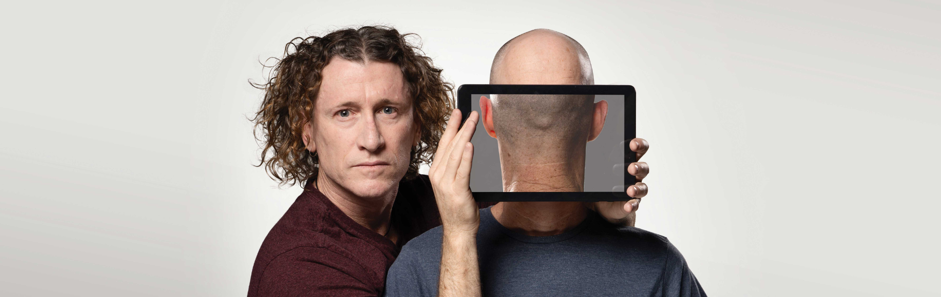 One man standing next to another holding a screen in front of the other's face. On the screen the image looks like the back of the man's head.