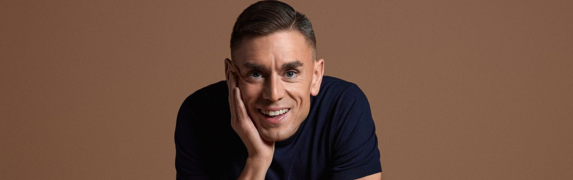 A close up of James Mullinger dressed in black with a brown background.