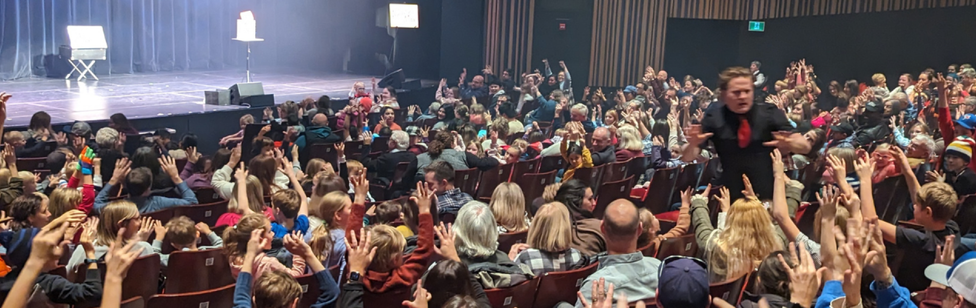 A full audience of children with their hands up in the air as the magician stands in the theatre aisle.