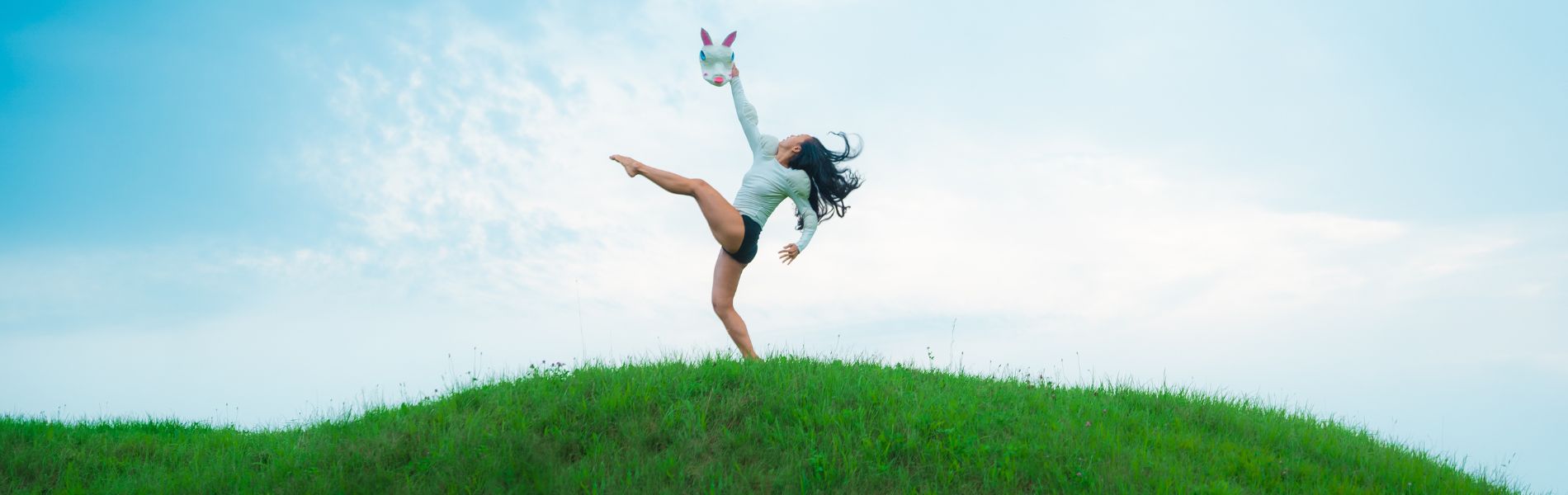 A dancer striking a post with limbs extended in front of a cloudy blue sky, on top of a bright green hill