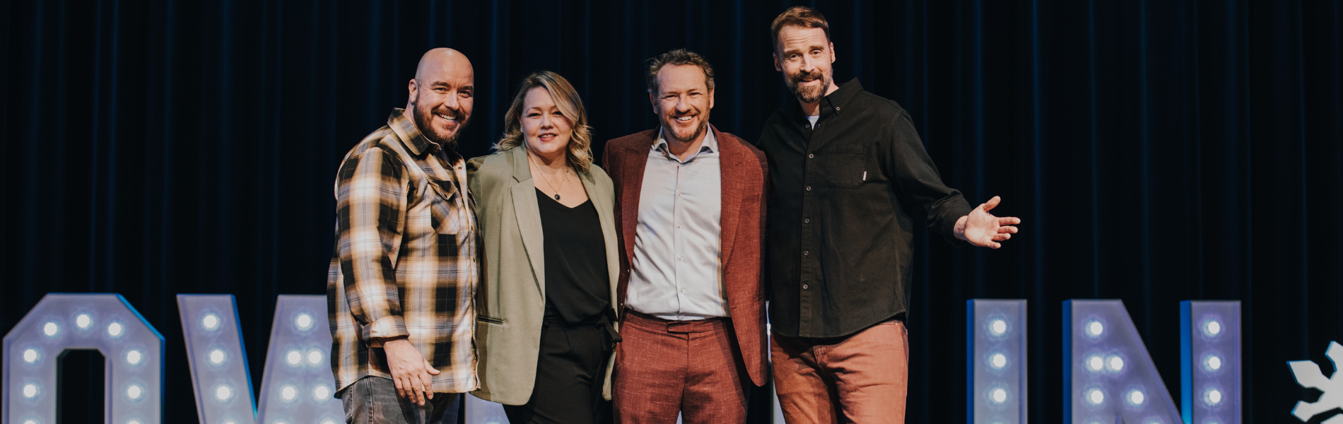 Four comedians posing on a stage in front of a lit sign.