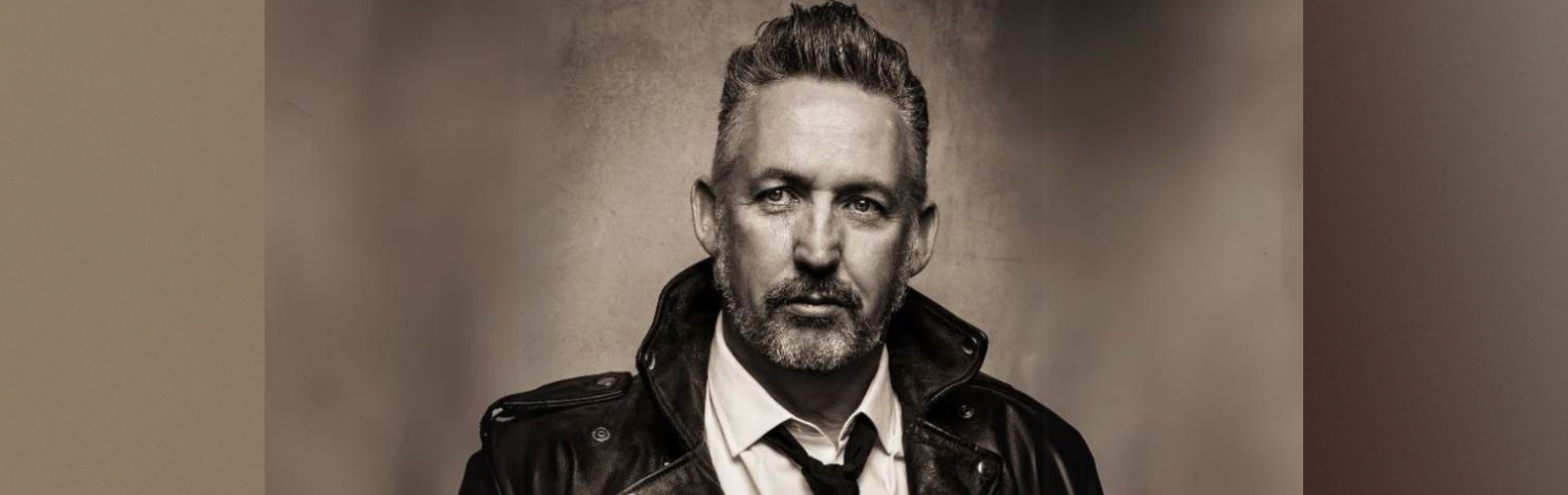 A sepia toned image of Harland Williams in a leather coat and tie.