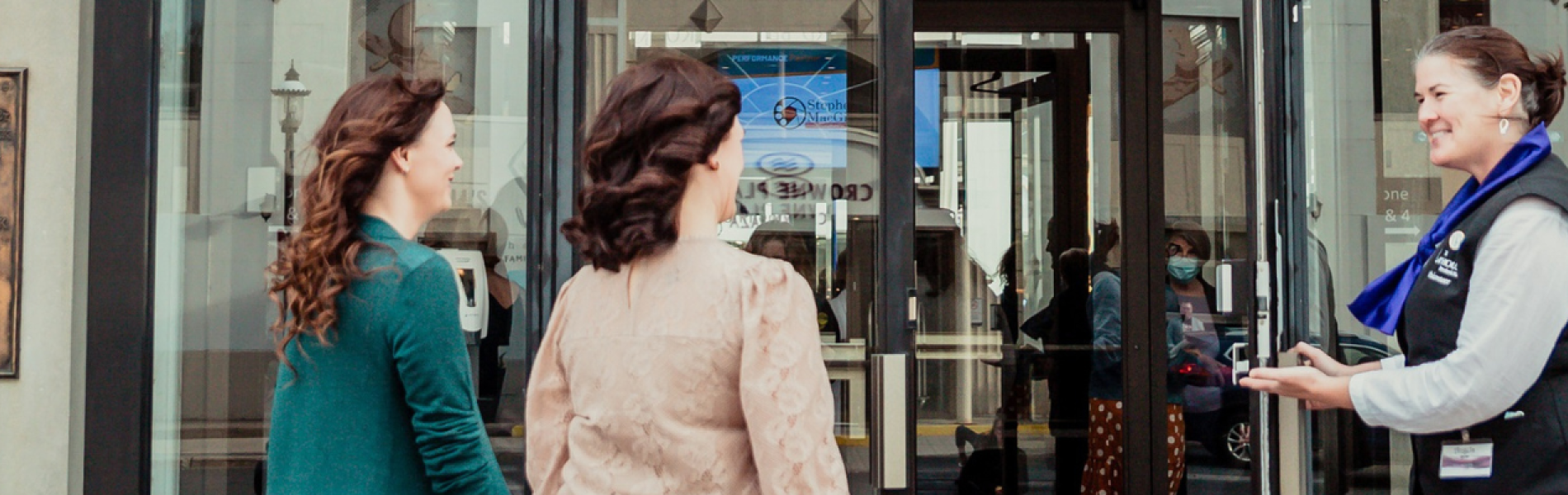 Two women with brown hair walking towards the front door of the Playhouse, held open by a welcoming and smiling volunteer usher 