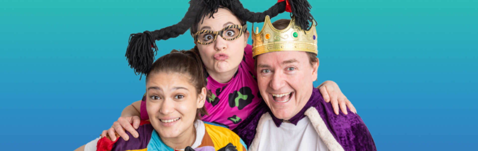 Three performers of DuffleBag Theatre huddled together in the centre of the photo, each are smiling and dressed in capes and costume pieces