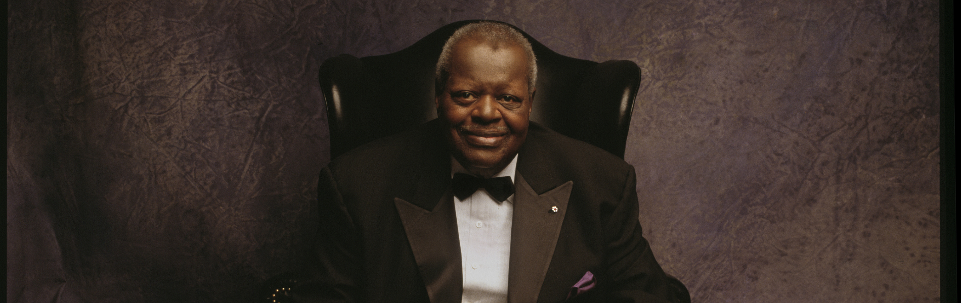 A closely cropped image of Oscar Peterson from the waist up, sitting in a high backed chair wearing a black tuxedo jacket, white shirt, and black botwie