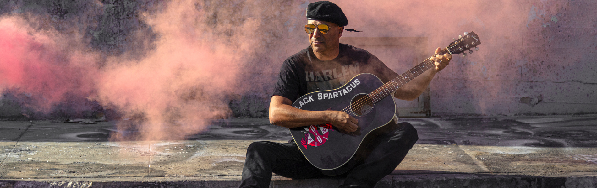 Tom Morello sitting on a sidewalk holding a black acoustic guitar in his lap. He's wearing reflective sunglasses, looking away from the camera, with pink smoke surrounding him