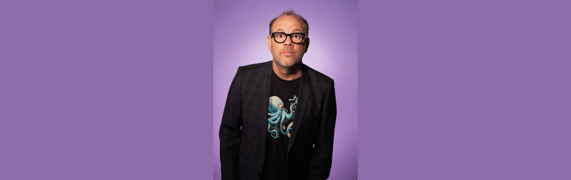 Tom Papa in a black blazer and black tshirt looking directly at the camera, standing in front of a light purple background