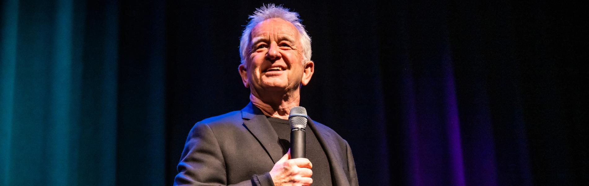 An image of Ron James on stage holding a microphone. 