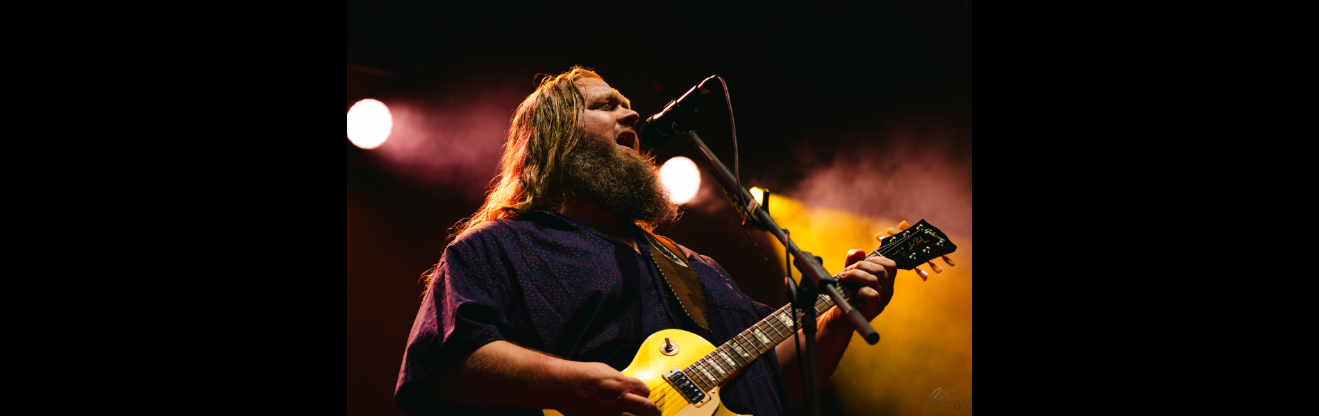 An image of Matt Andersen singing into a mic while playing guitar with pink and yellow lighting behind. 