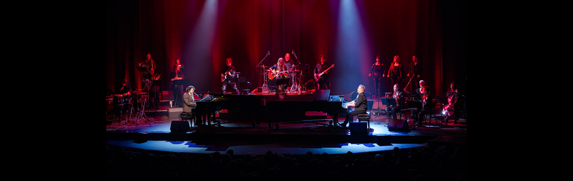 Two grand pianos and pianists in the foreground with a band lit in red surrounding them. 