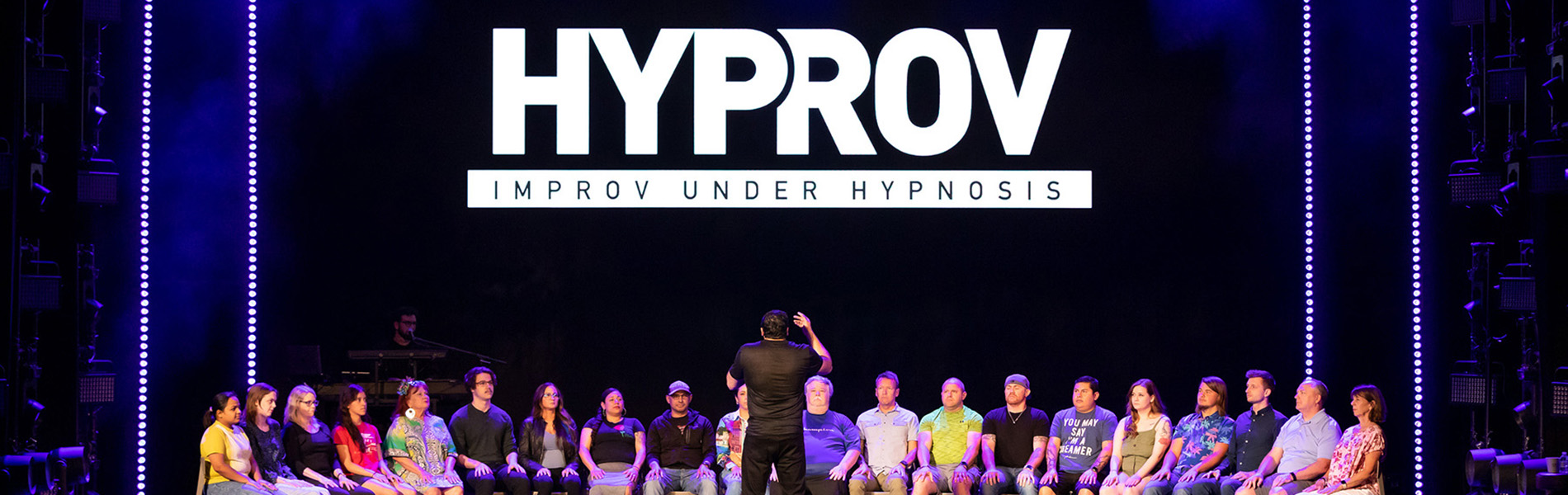 A group of people in chairs on the stage with the words, 'HYPROV Improv under hypnosis' in white lettering above them.