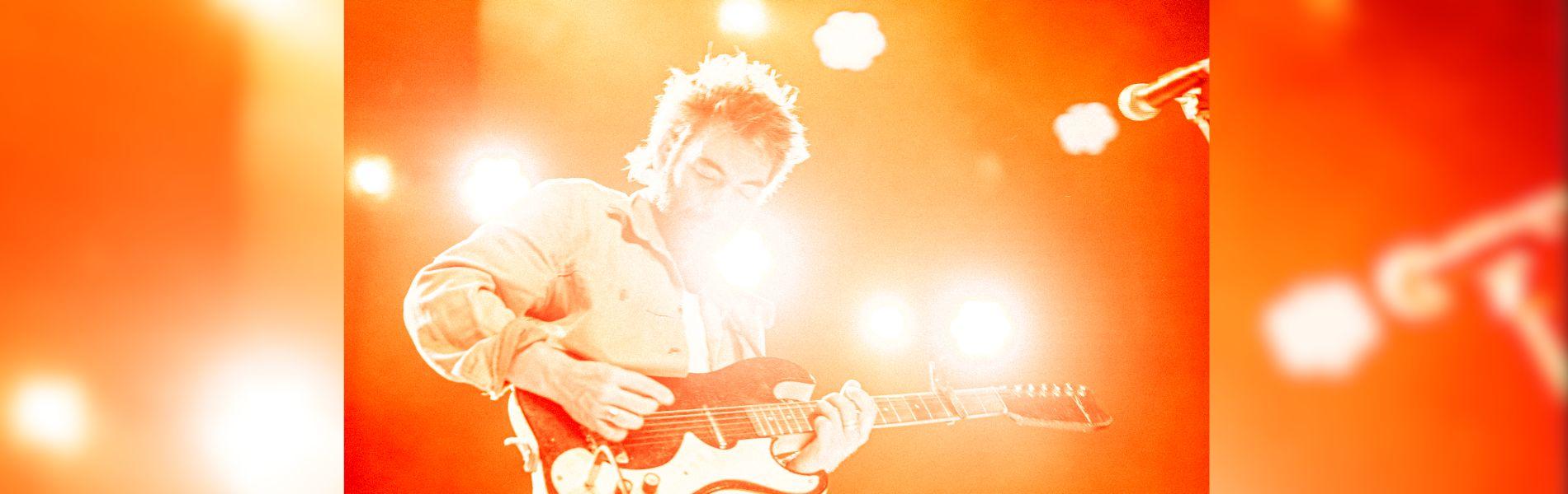 Leif stands on a smoky, colourful stage holding a guitar.