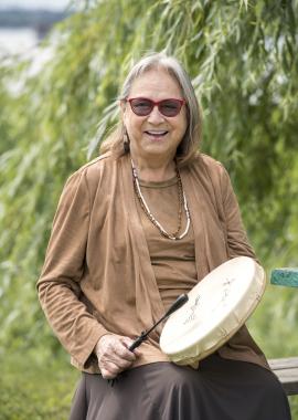 Elder Maggie Paul holding a drum in front of trees