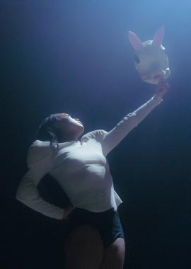 A dancer stands under a spotlight holding a white rabbit mask up in the air.